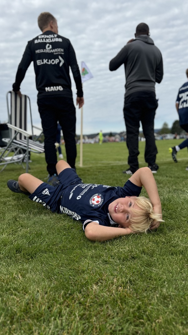 Fra sidelinjen på en fotballkamp, ryggen til en trener og en tilskuer, ung fotballspiller som ligger i gresset og ser i kameraet.